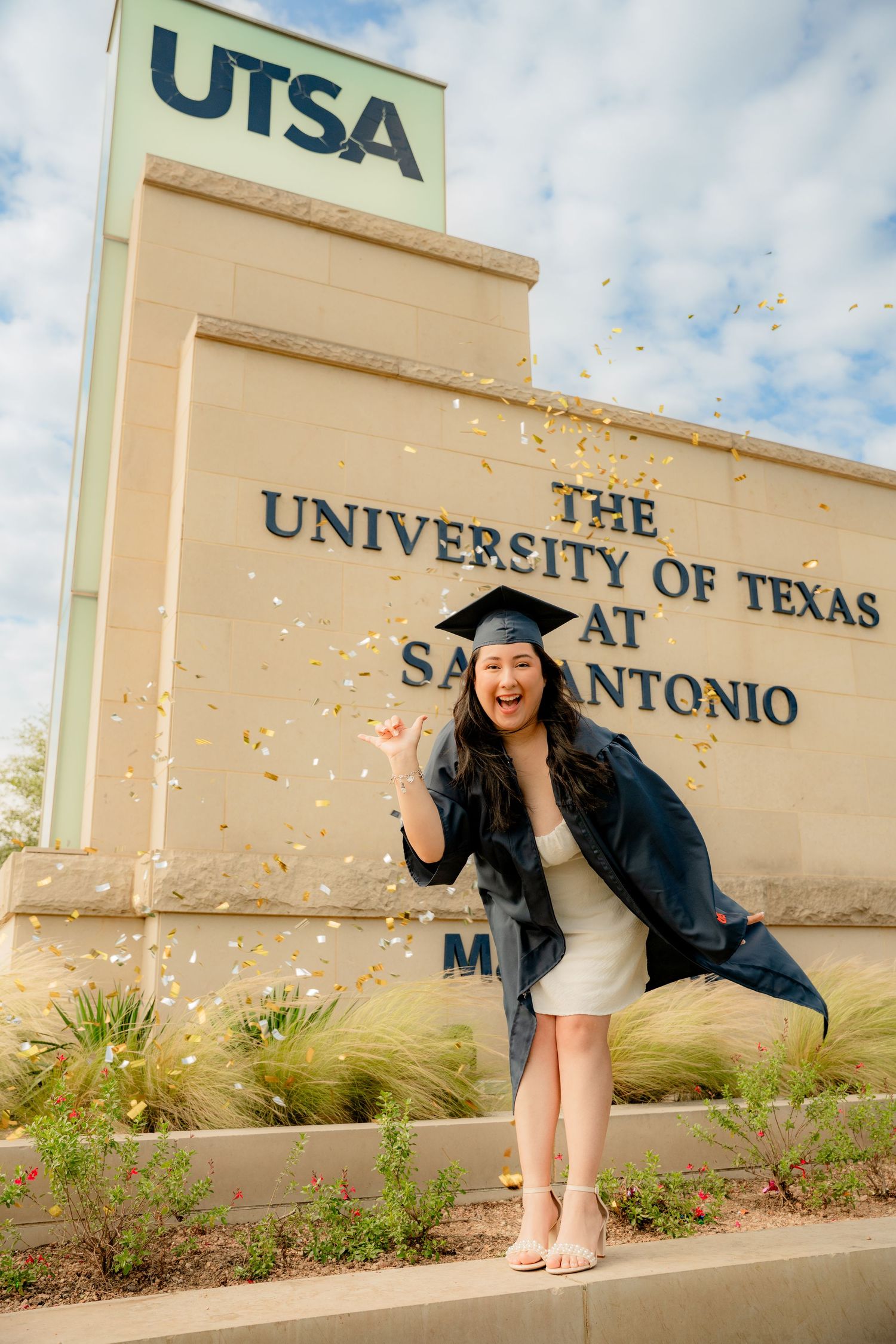 Graduate in cap and gown celebrates with confetti in front of UTSA sign.