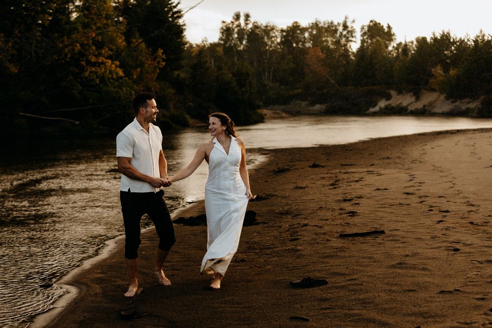 Couple strolling on a dark sandy beach with trees in background.