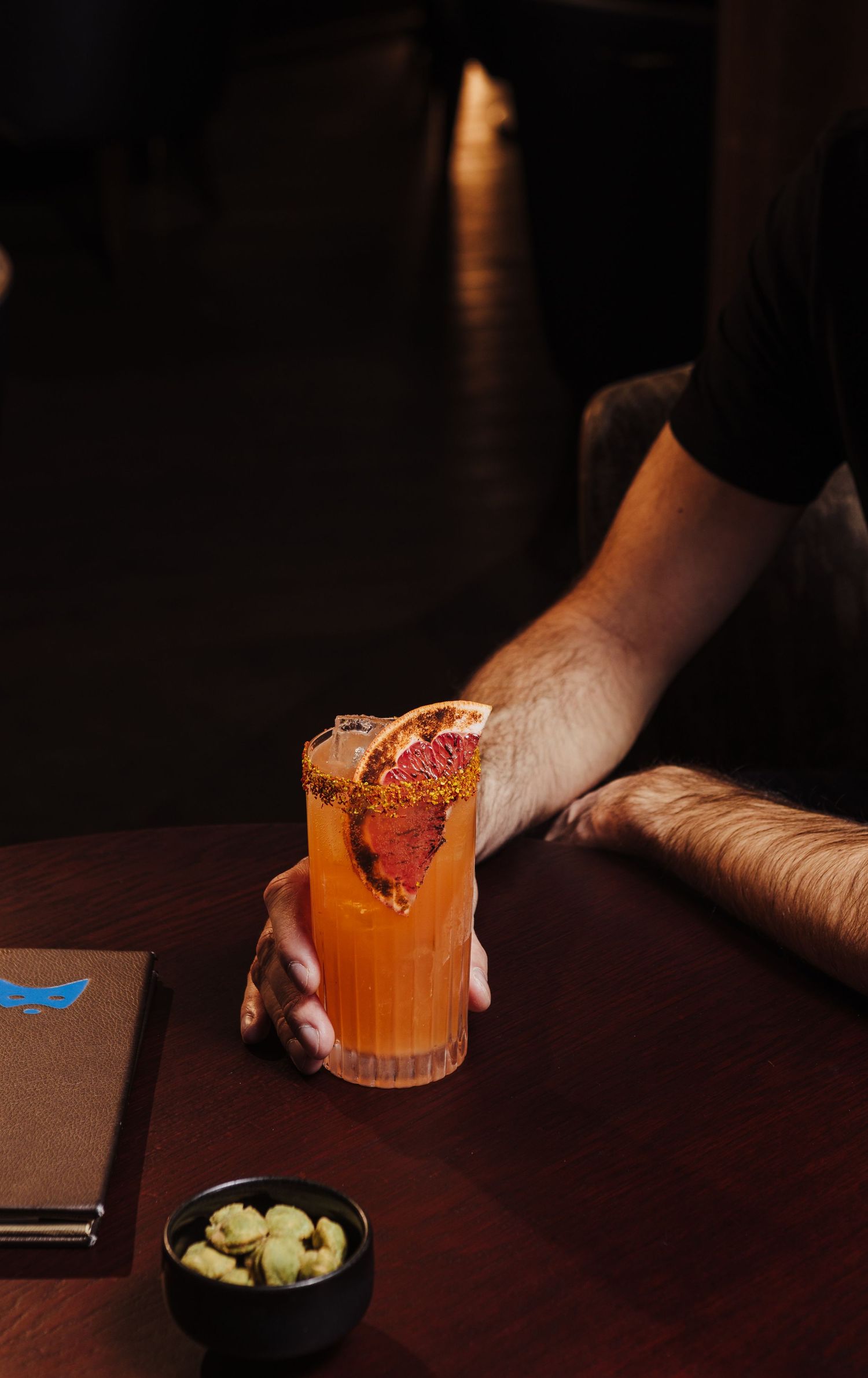 Orange cocktail with garnish being served on dark wooden bar top next to small bowl of snacks.