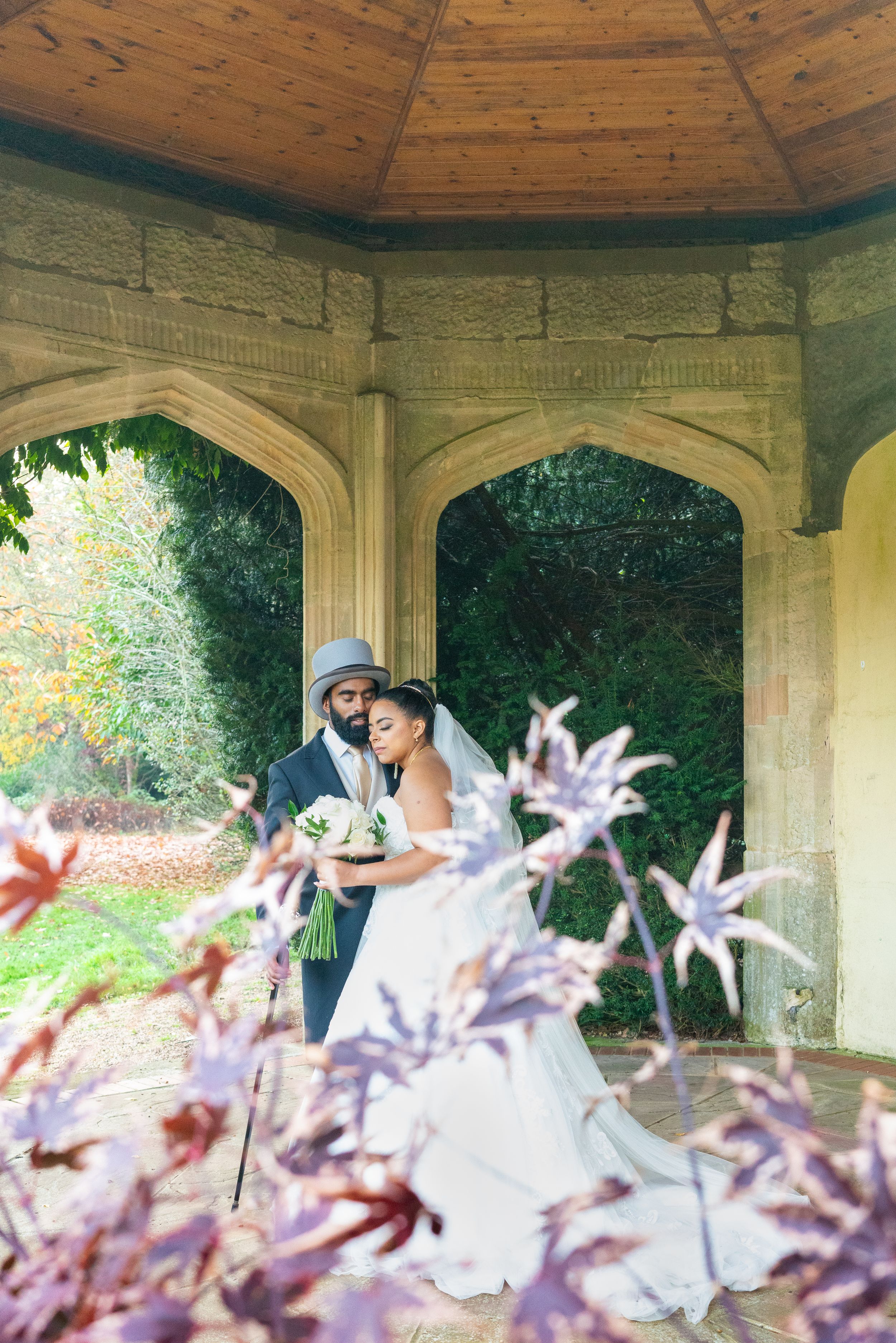 A romantic wedding couple embraces in an elegant stone gazebo surrounded by soft purple flowers and greenery.