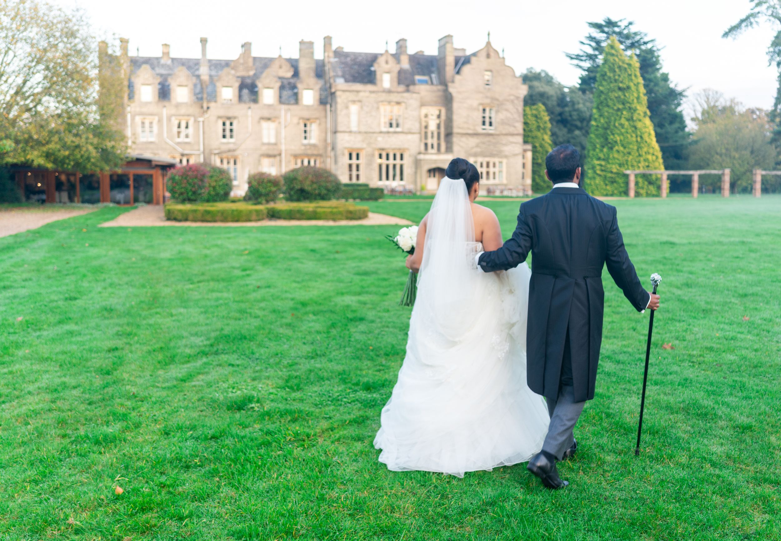 A couple in formal wedding attire walks across a manicured lawn toward a grand historic stone castle mansion.