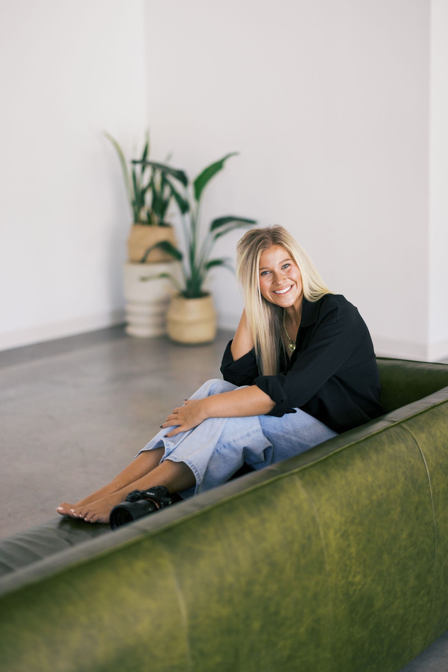 Casual portrait of woman in black sweater relaxing on green velvet couch with plants in background.