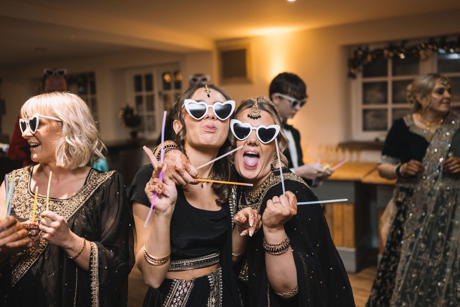 Party guests wearing fun novelty sunglasses pose with peace signs while celebrating during a festive indoor gathering.