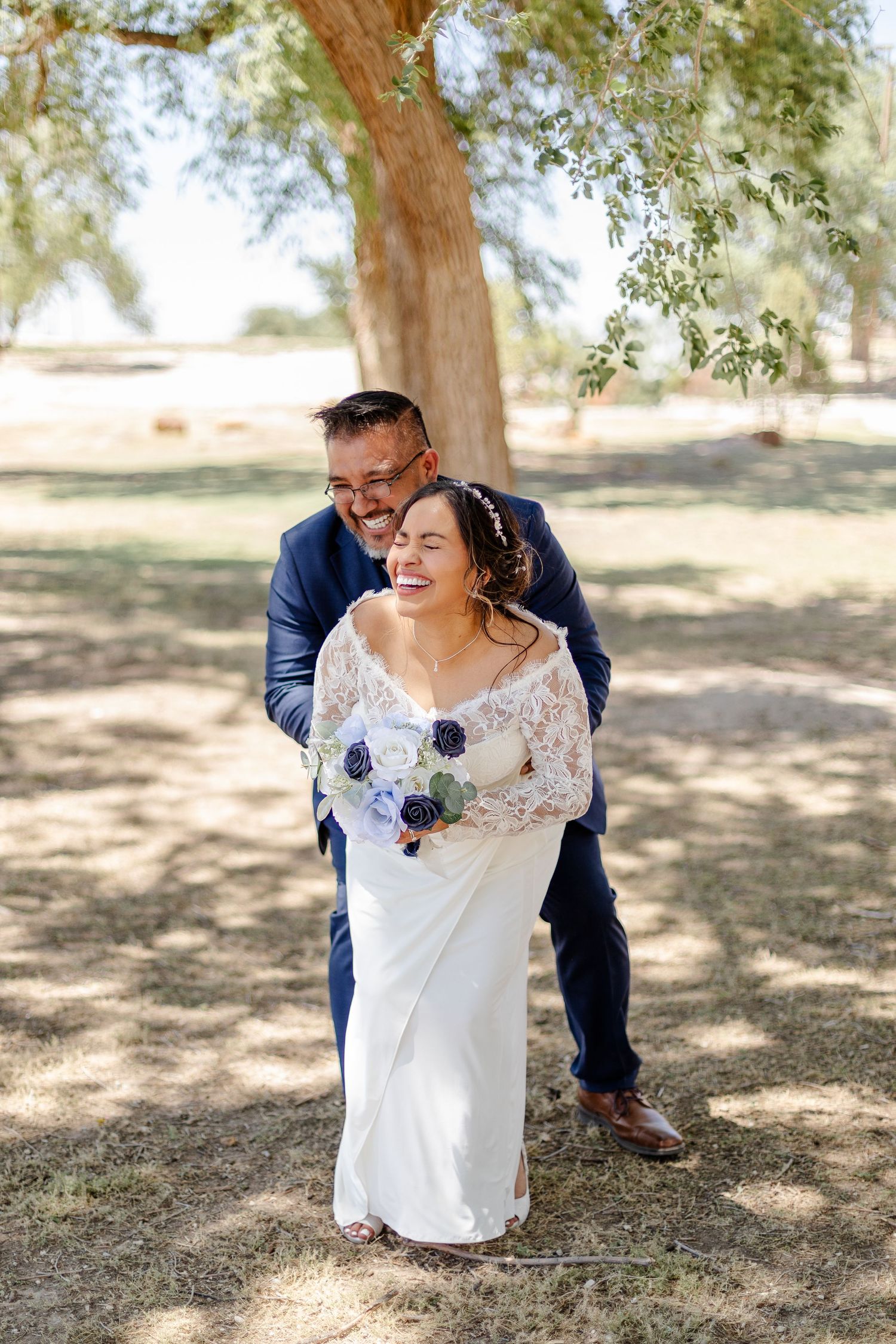 A joyful wedding moment captured under a tree with a bride in lace holding a blue bouquet.