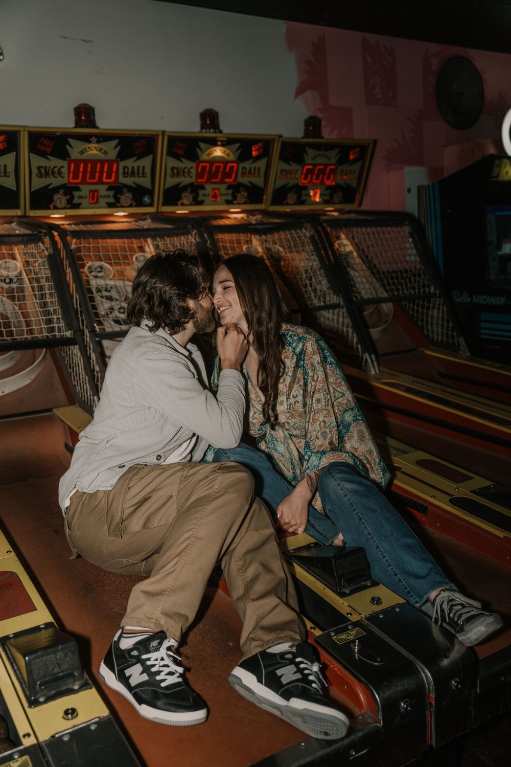 Two people relax on arcade bowling alley benches in casual attire under neon lighting.