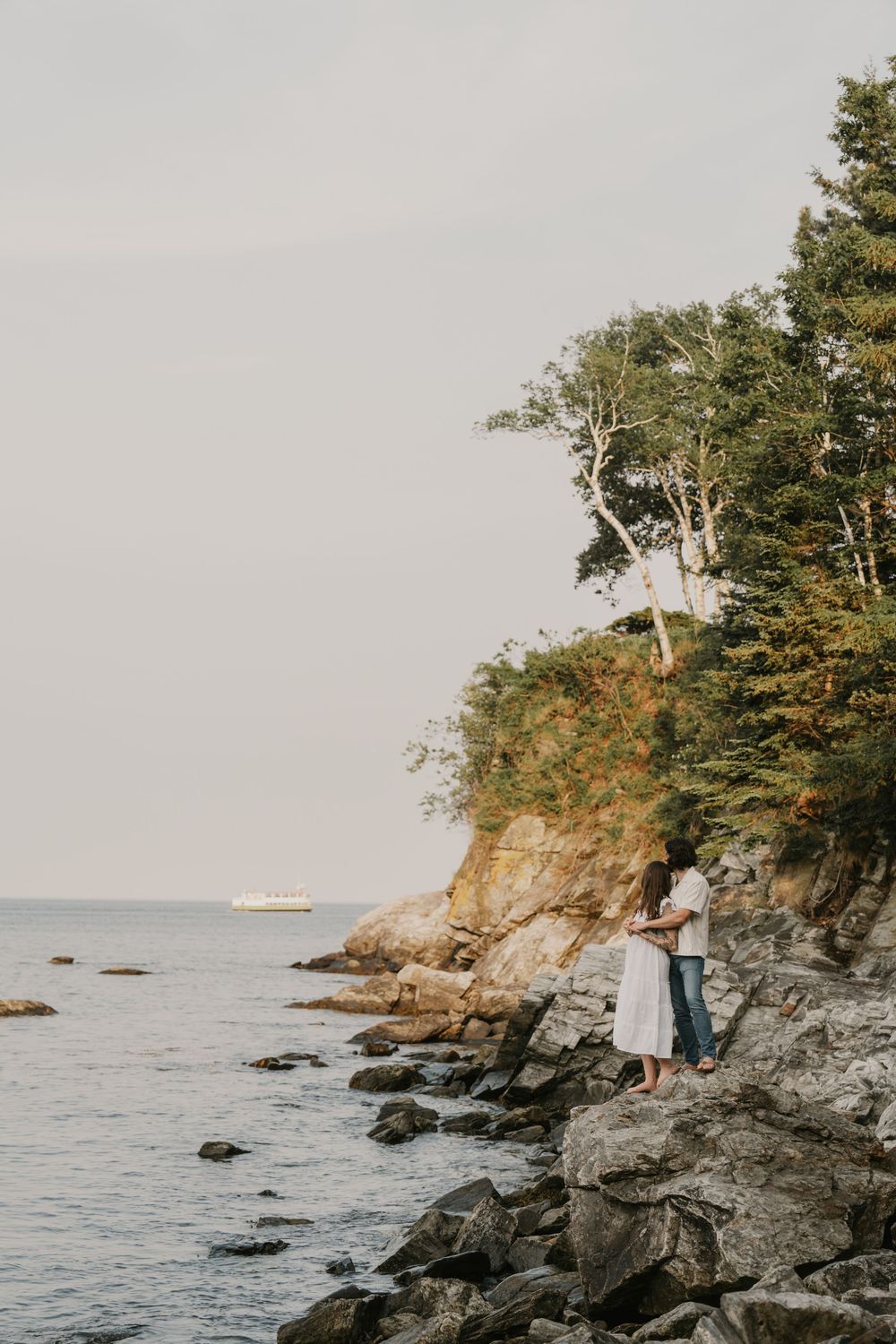 A couple stands on rocky coastal cliffs overlooking the ocean at sunset with evergreen trees on the shoreline.