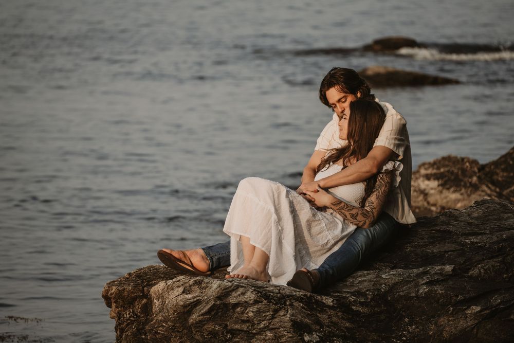 Figure in white dress sits contemplatively on rocky shore at sunset by tranquil water.