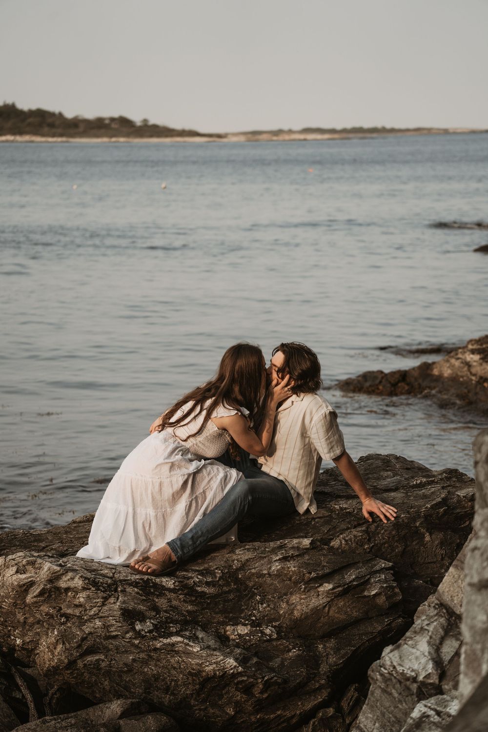 A romantic couple shares an intimate moment on coastal rocks by the ocean at sunset.