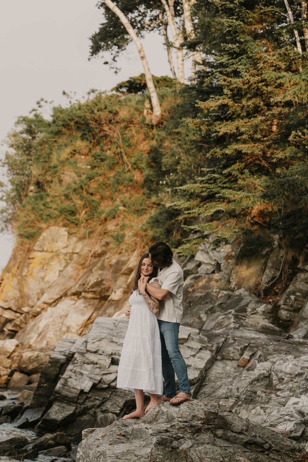 A couple sits together on rocky coastal cliffs during sunset as waves crash below on the Pacific Northwest shore.