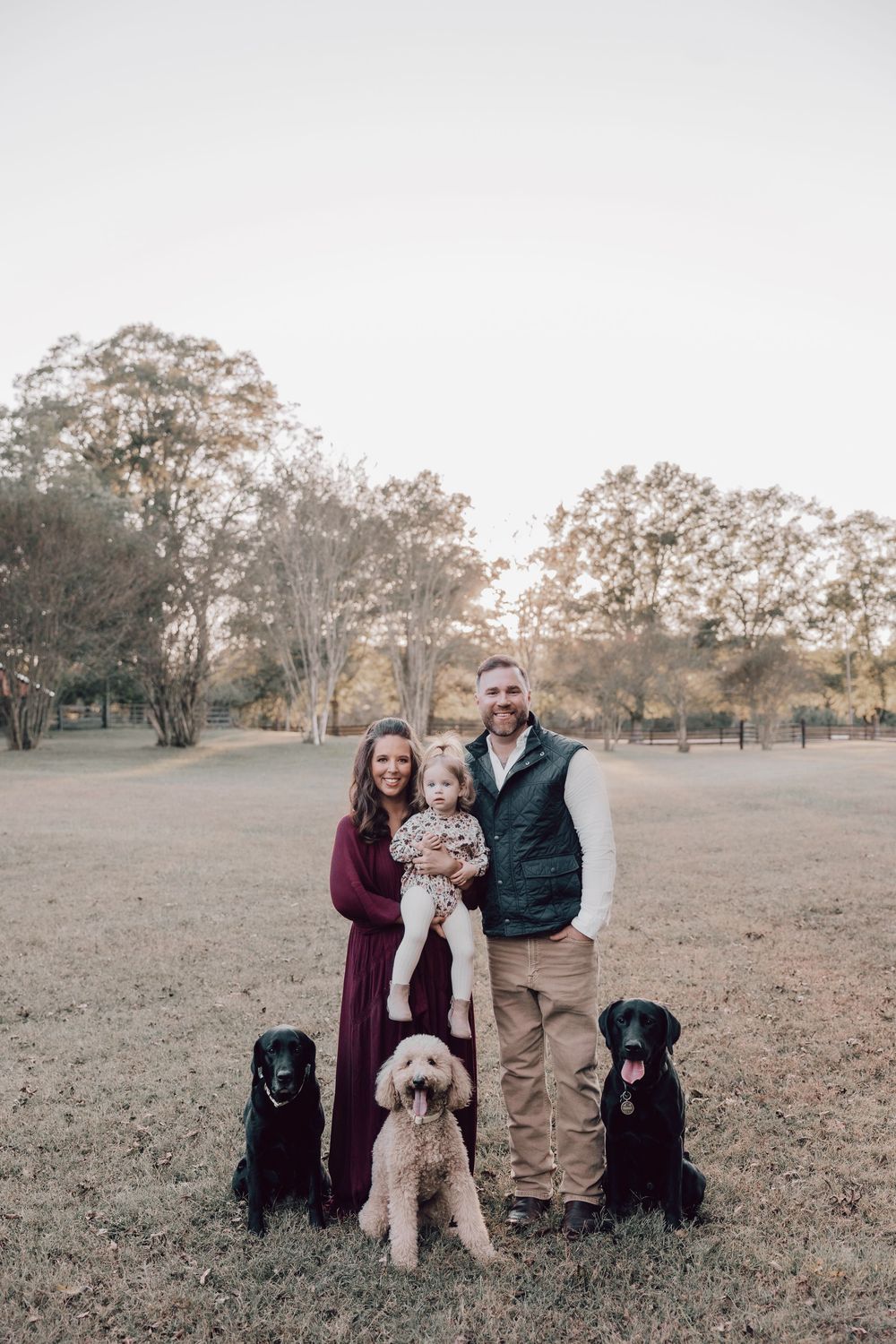 A family poses together with their black Labrador dogs in an autumn field at sunset.