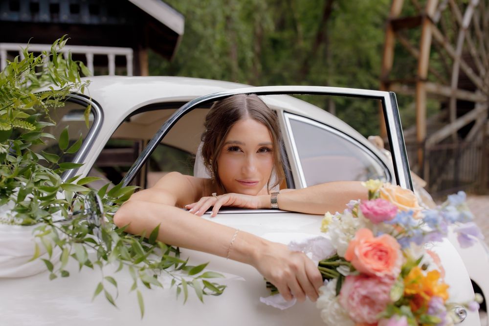 Bride leaning out of a vintage white car window, adorned with colorful floral arrangements on a wedding day.
