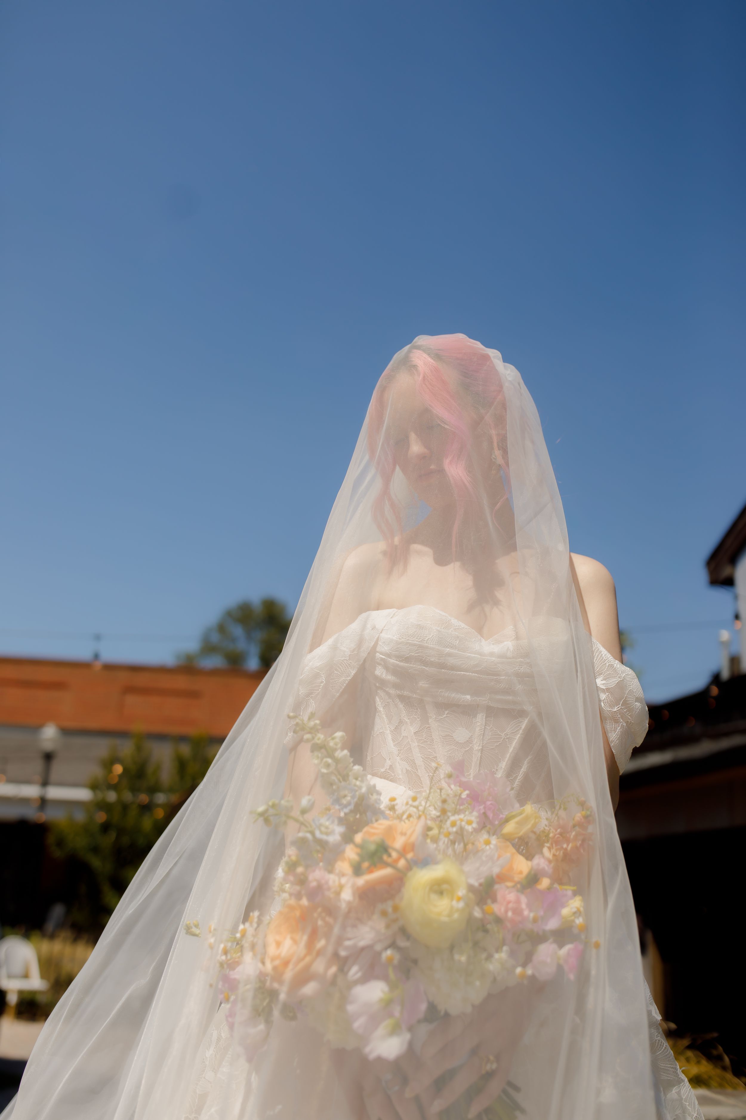 Bride in white gown and veil holding pastel bouquet outdoors under clear blue sky.