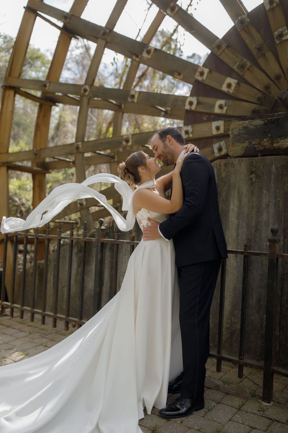 Bride and groom embracing under rustic wooden arch, wedding veil flowing in wind, outdoor ceremony venue.