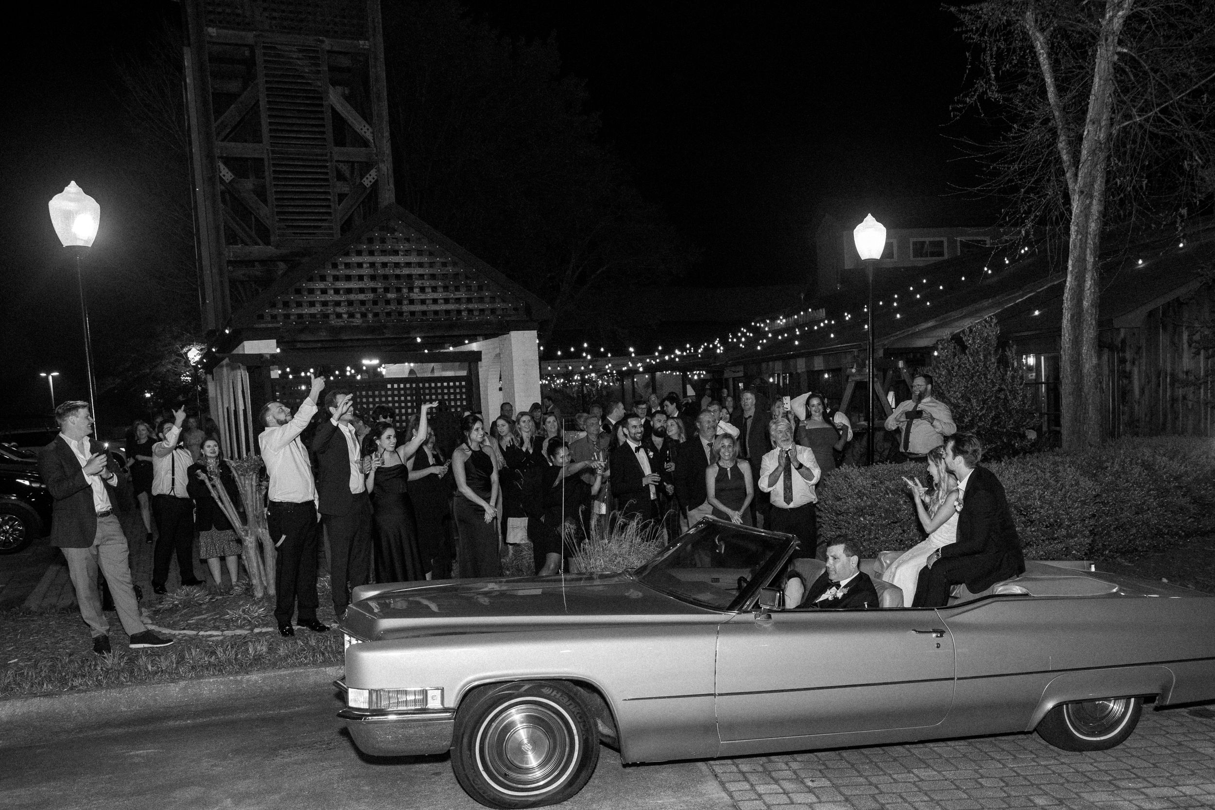 Wedding guests celebrate around a classic convertible car at night with street lamps glowing.