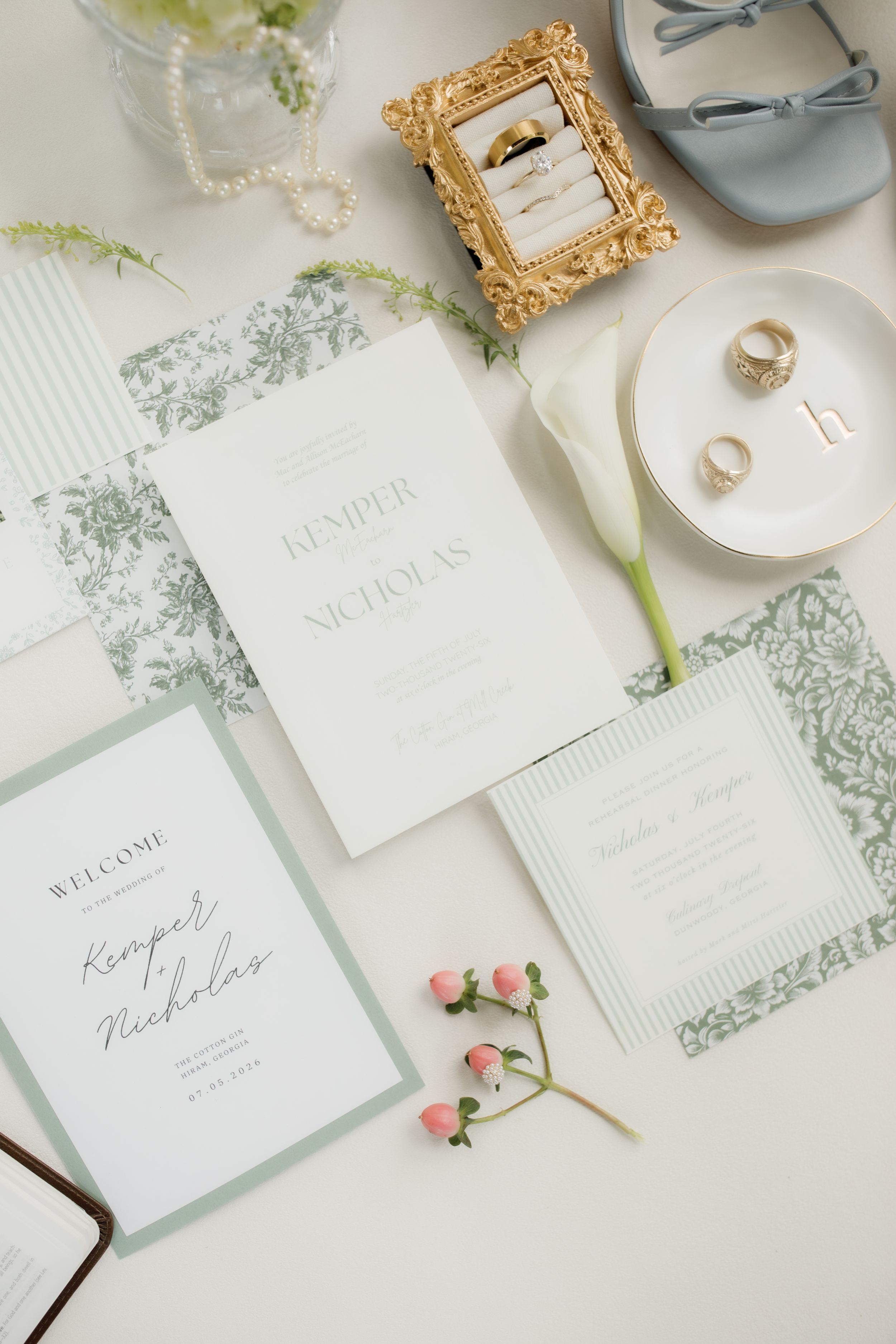 Elegant wedding stationery flat lay with sage green floral details, gold rings, pearl necklace, and calla lily accents.