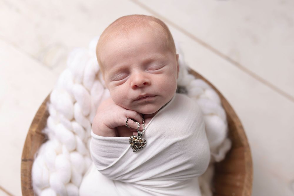 A sleeping newborn baby swaddled in white fabric lies peacefully in a wooden bowl with soft white accessories.