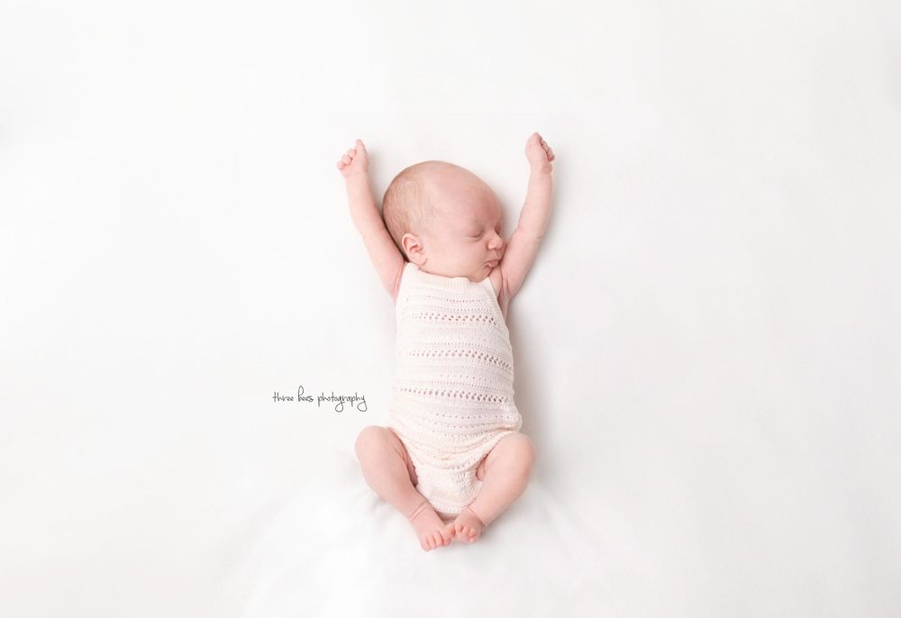 A newborn baby lying on a white background wearing soft pink clothing with arms stretched overhead.