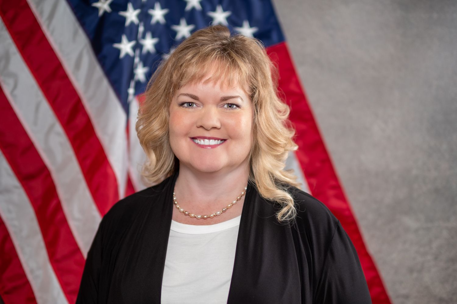 Professional portrait photo of a person with blonde hair wearing a black blazer and pearl necklace in front of an American flag.