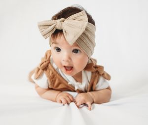 Adorable baby girl wearing a tan bow headband and brown romper, lying on white background with a surprised expression.