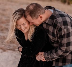 A smiling couple shares a tender embrace outdoors, with the man nuzzling the woman in a plaid shirt and black velvet dress.