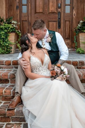 Bride and groom share a romantic kiss on brick steps before elegant wooden doors, bride holding a bouquet.