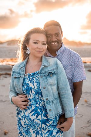 Couple posing on beach at golden sunset, woman in blue floral dress and denim jacket, man in patterned shirt.