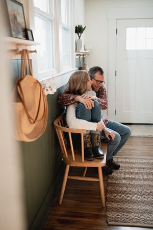 A father and daughter share a warm embrace while sitting in a wooden chair in a cozy home entryway.
