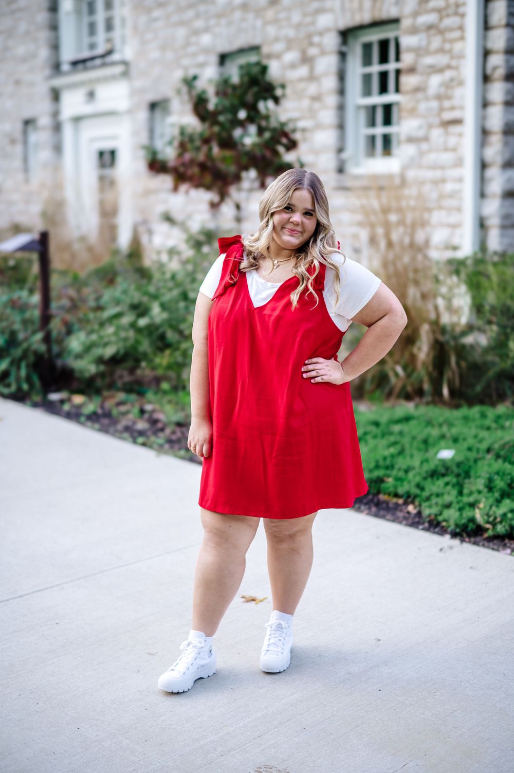 Person in red dress and white sneakers poses confidently against stone building backdrop.