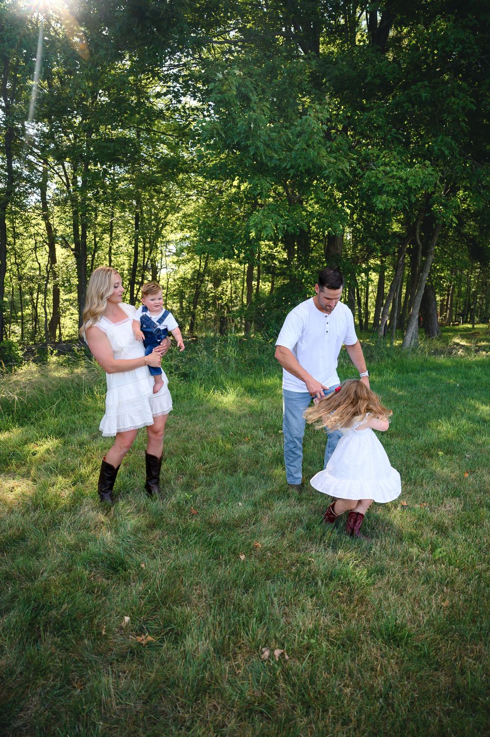 Family of four playing outdoors in lush green field, mom holding baby boy while dad twirls daughter in white dress.