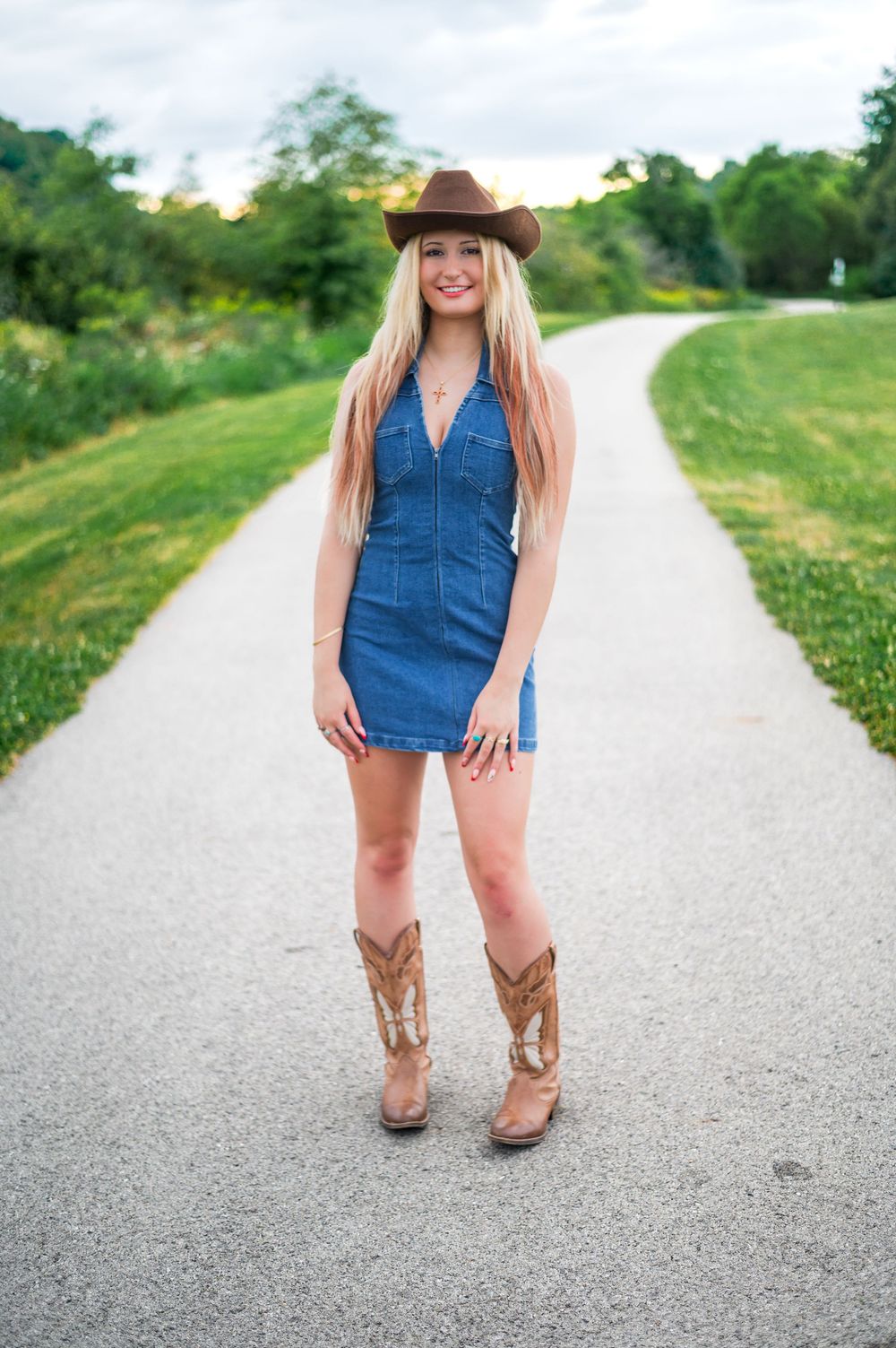 Person in denim dress and brown cowboy boots poses on gravel country road.
