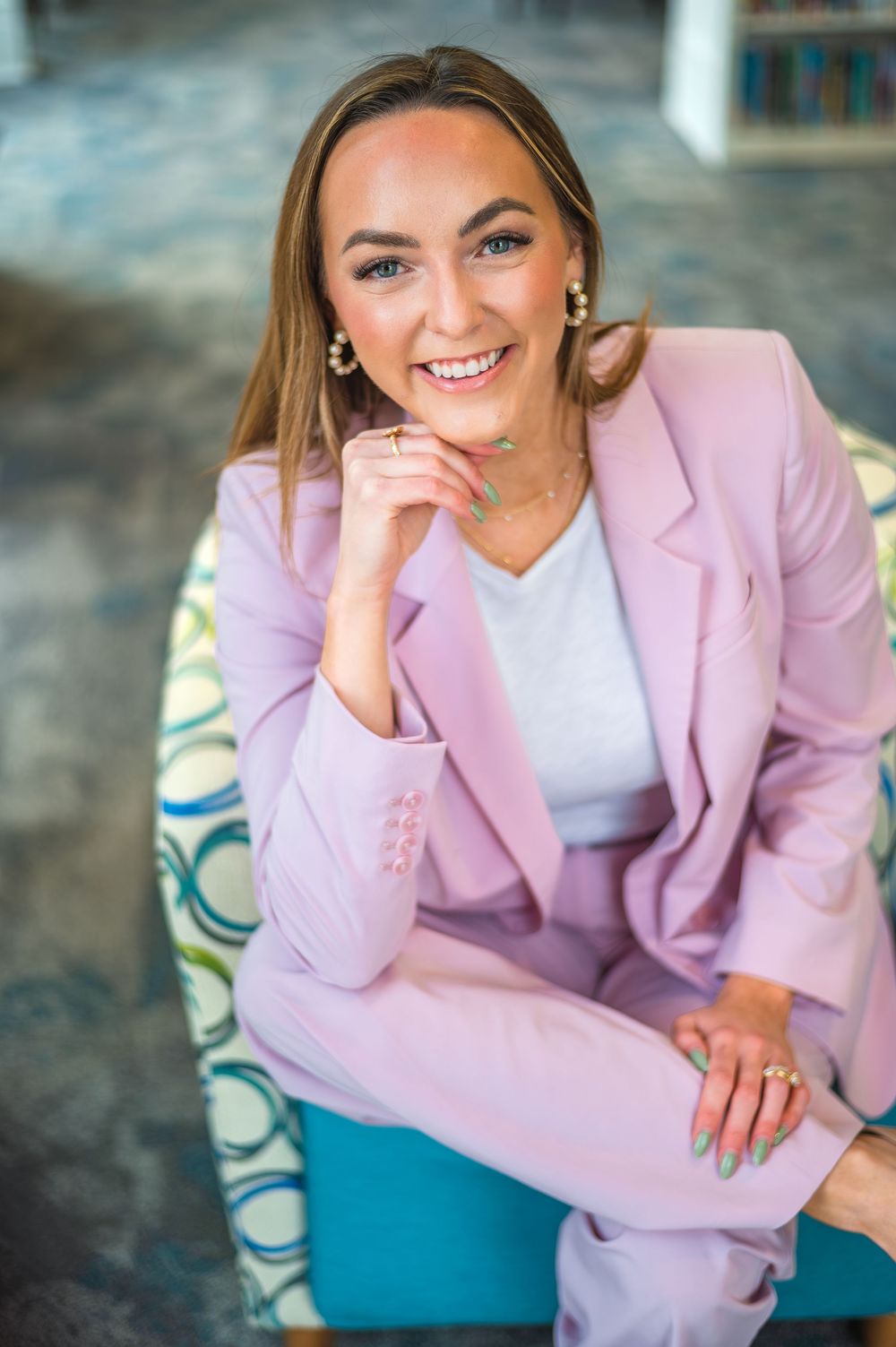 Woman in pink blazer and white top poses casually on blue patterned chair.
