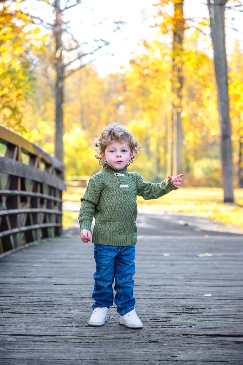 A small child in a green sweater and jeans stands on a wooden bridge during golden autumn.