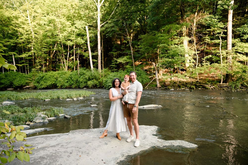 Family of three posing on rocks in a serene forest creek surrounded by lush green trees.
