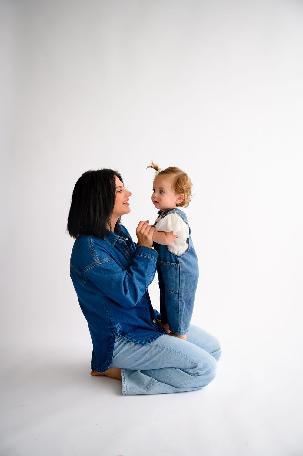 A mother in denim sits on white floor holding her smiling toddler in denim overalls, sharing a joyful moment.