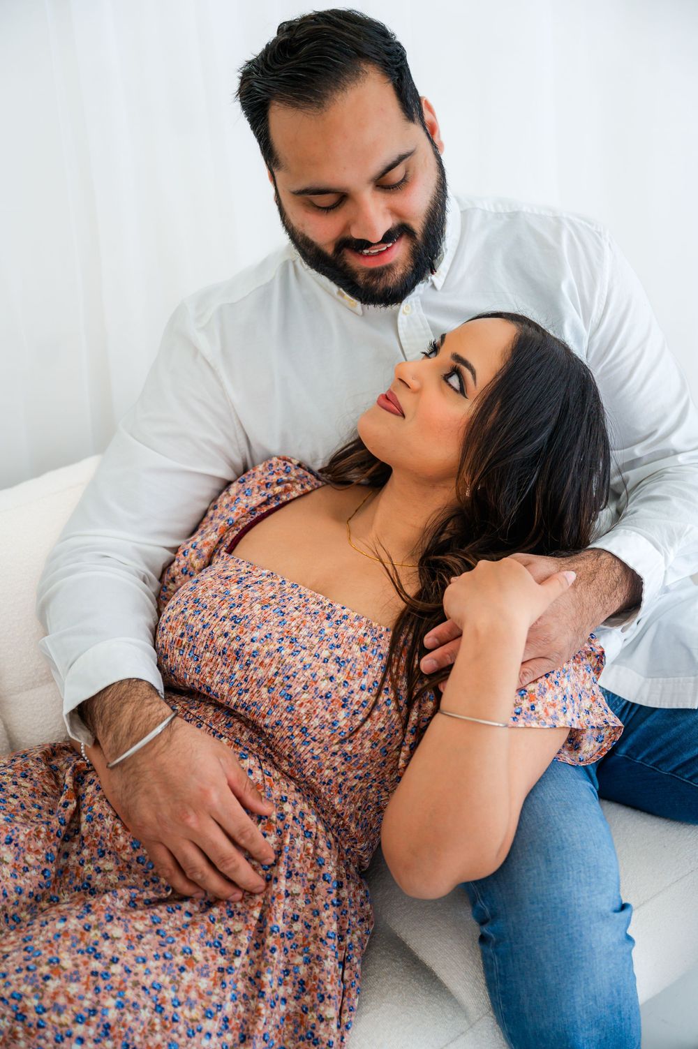 Couple in casual attire sharing intimate moment on white couch during indoor photoshoot.