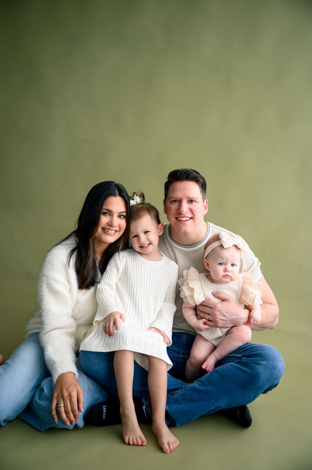 A happy family of four poses together on a green backdrop, with two young daughters in white outfits and parents in casual attire.