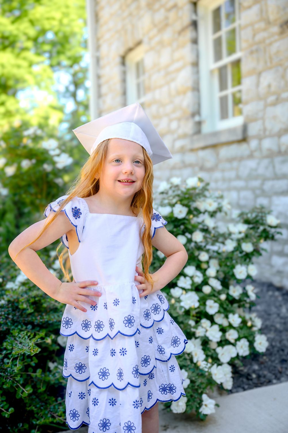 Child in white eyelet dress and bonnet posing by stone building with blooming flowers.