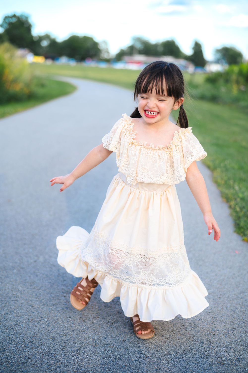 A child in a flowing white lace dress twirls and dances along a paved rural road on a sunny day.