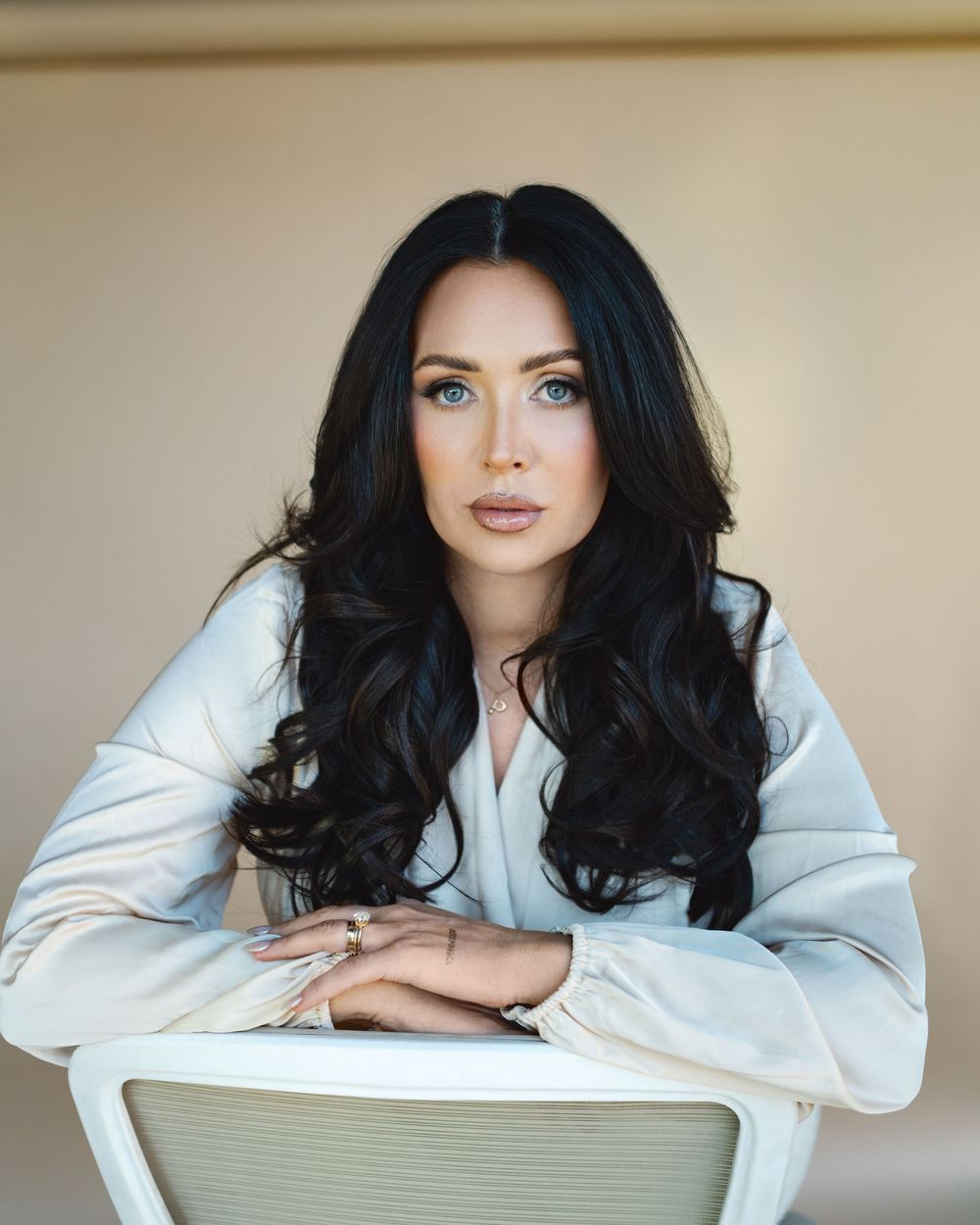 Portrait of a woman with long dark hair wearing a light colored top leaning on a white chair against a neutral background.