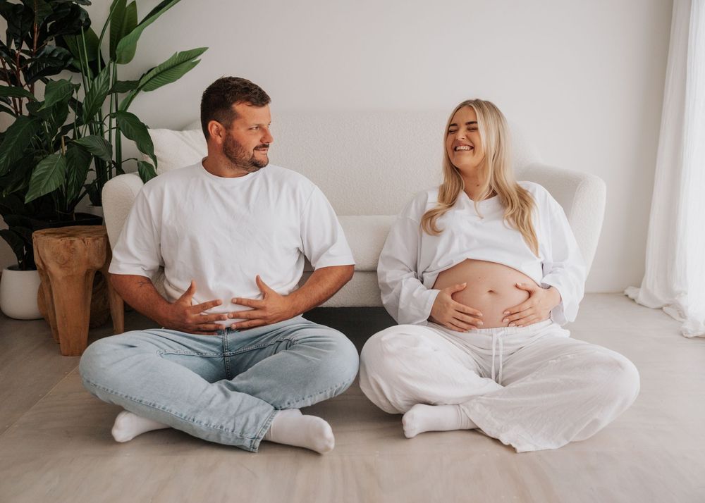 Pregnant couple sitting on floor in white outfits, man looking at woman who smiles while cradling her baby bump.