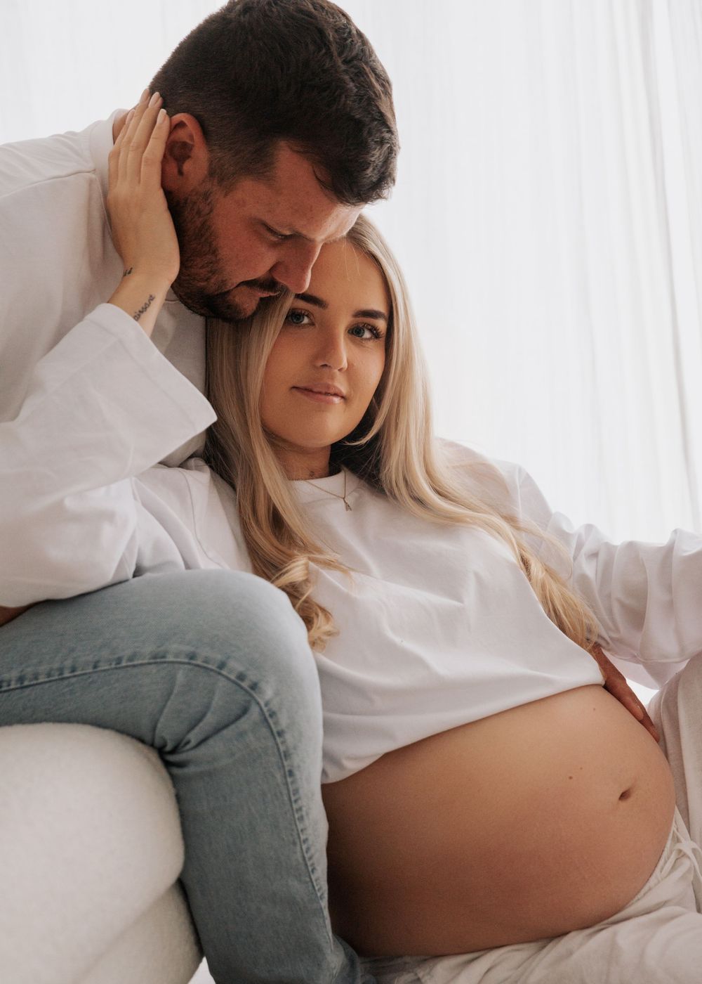 A pregnant woman in a white top sits with her partner, both sharing a tender intimate moment together.