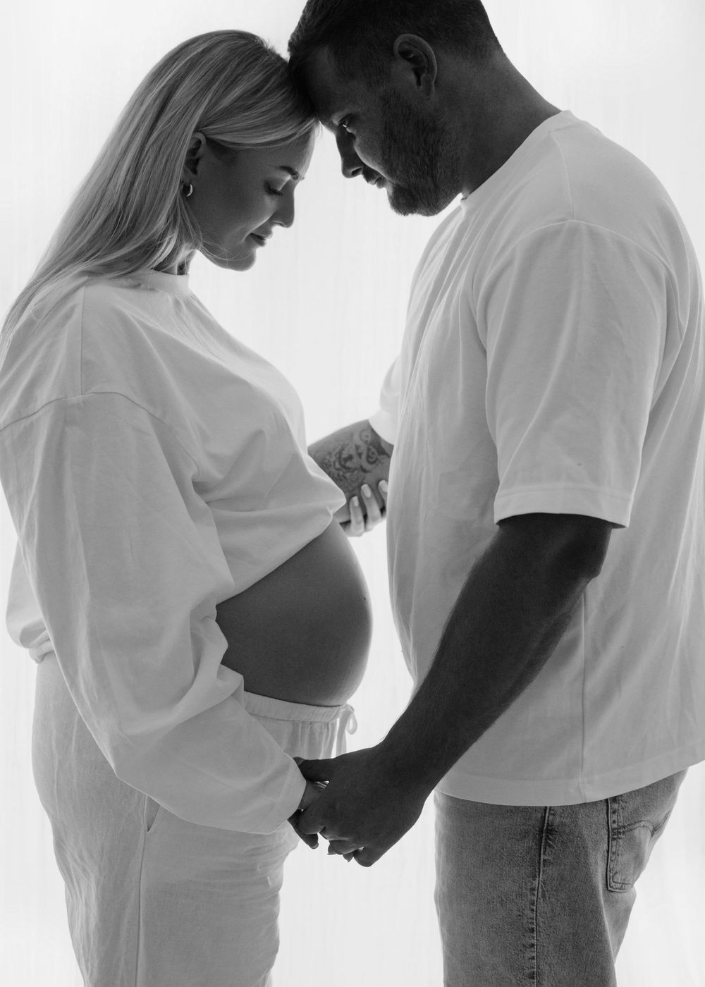 Black and white maternity photo of a pregnant couple holding hands, touching foreheads tenderly in white outfits.