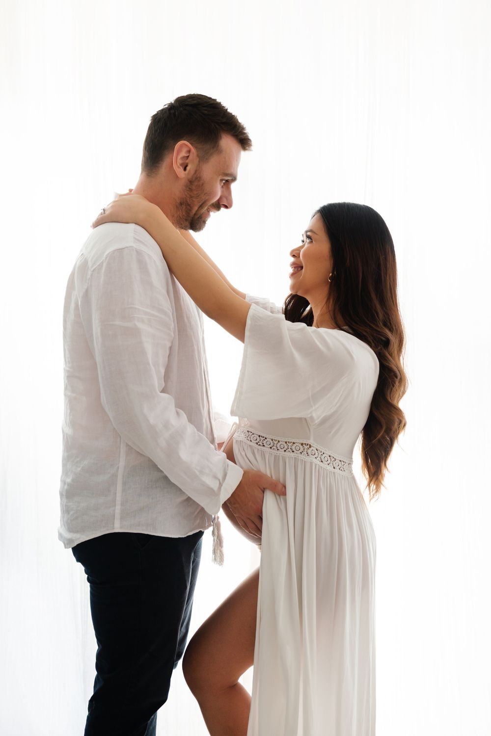 A happy couple in white outfits share a tender moment, with the pregnant woman smiling at her partner.