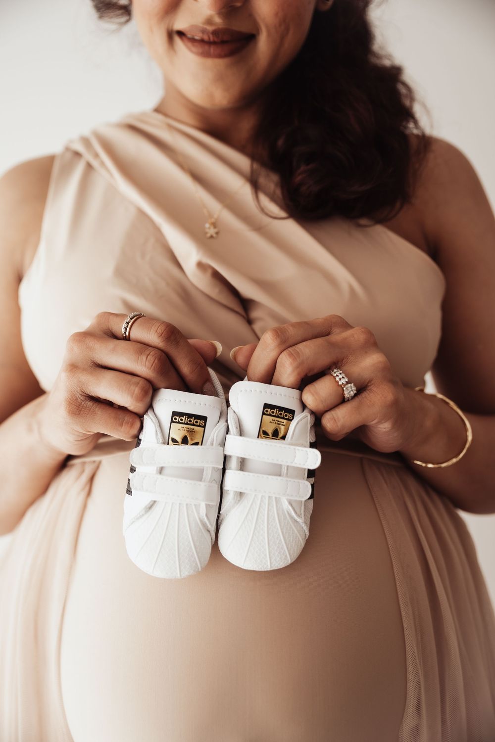 Pregnant woman in beige dress holding tiny white Adidas Superstar baby sneakers over her baby bump.