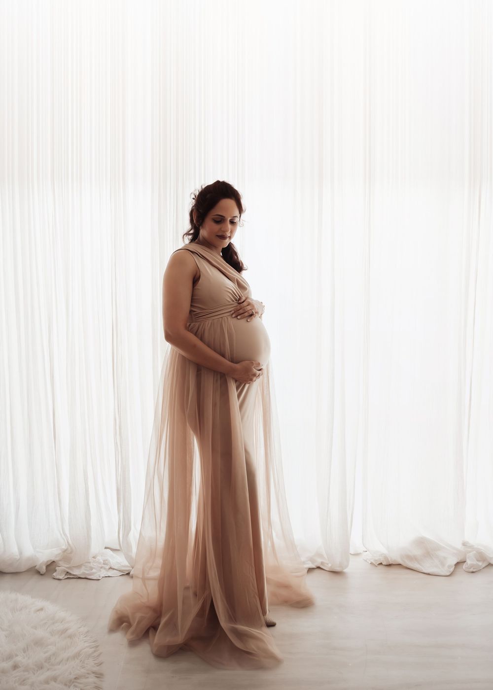 Pregnant woman in elegant beige tulle maternity gown cradling baby bump, standing before sheer white curtains.