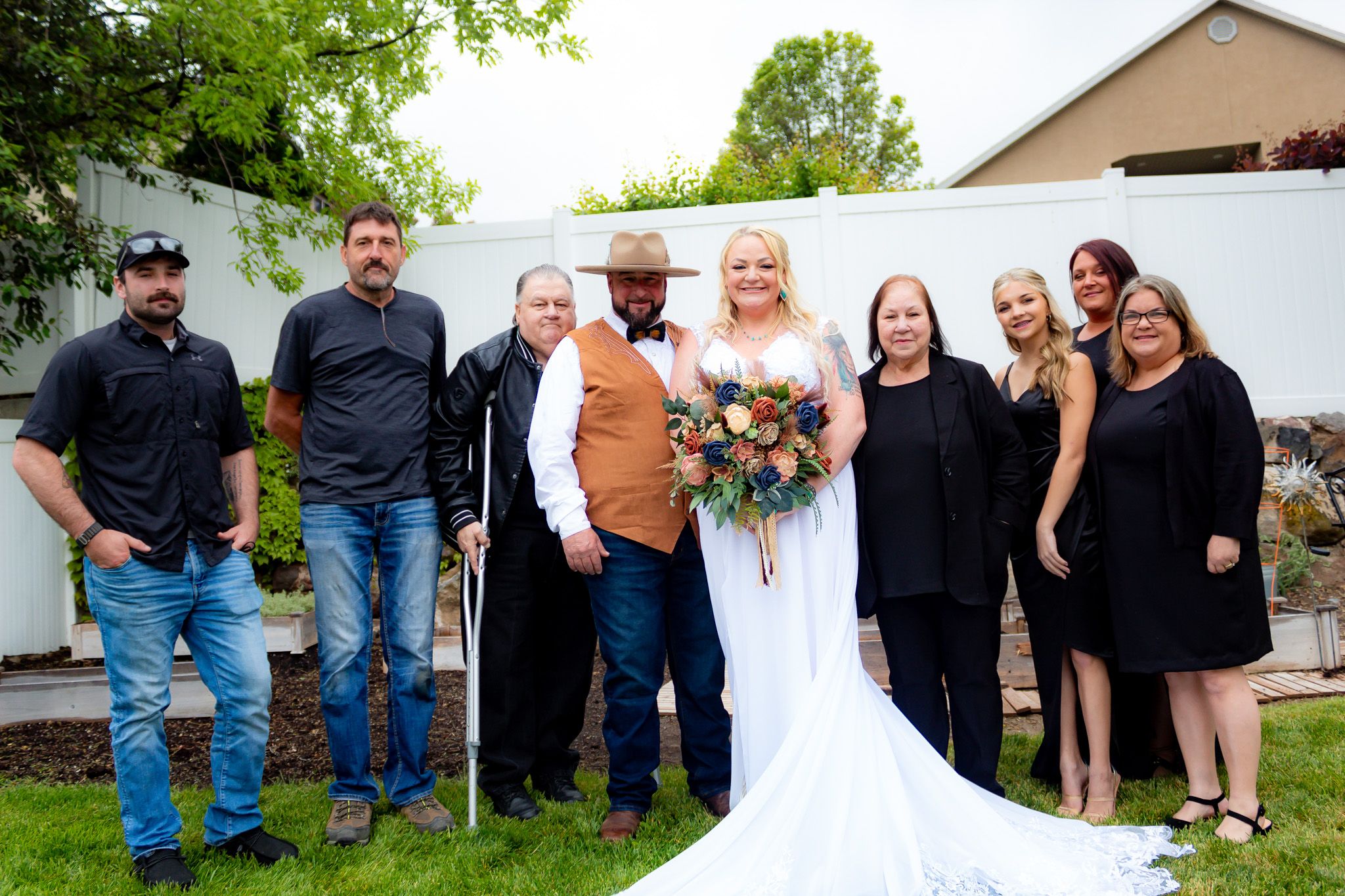 Wedding party poses together on a lawn with white fence backdrop and beautiful floral bouquet.