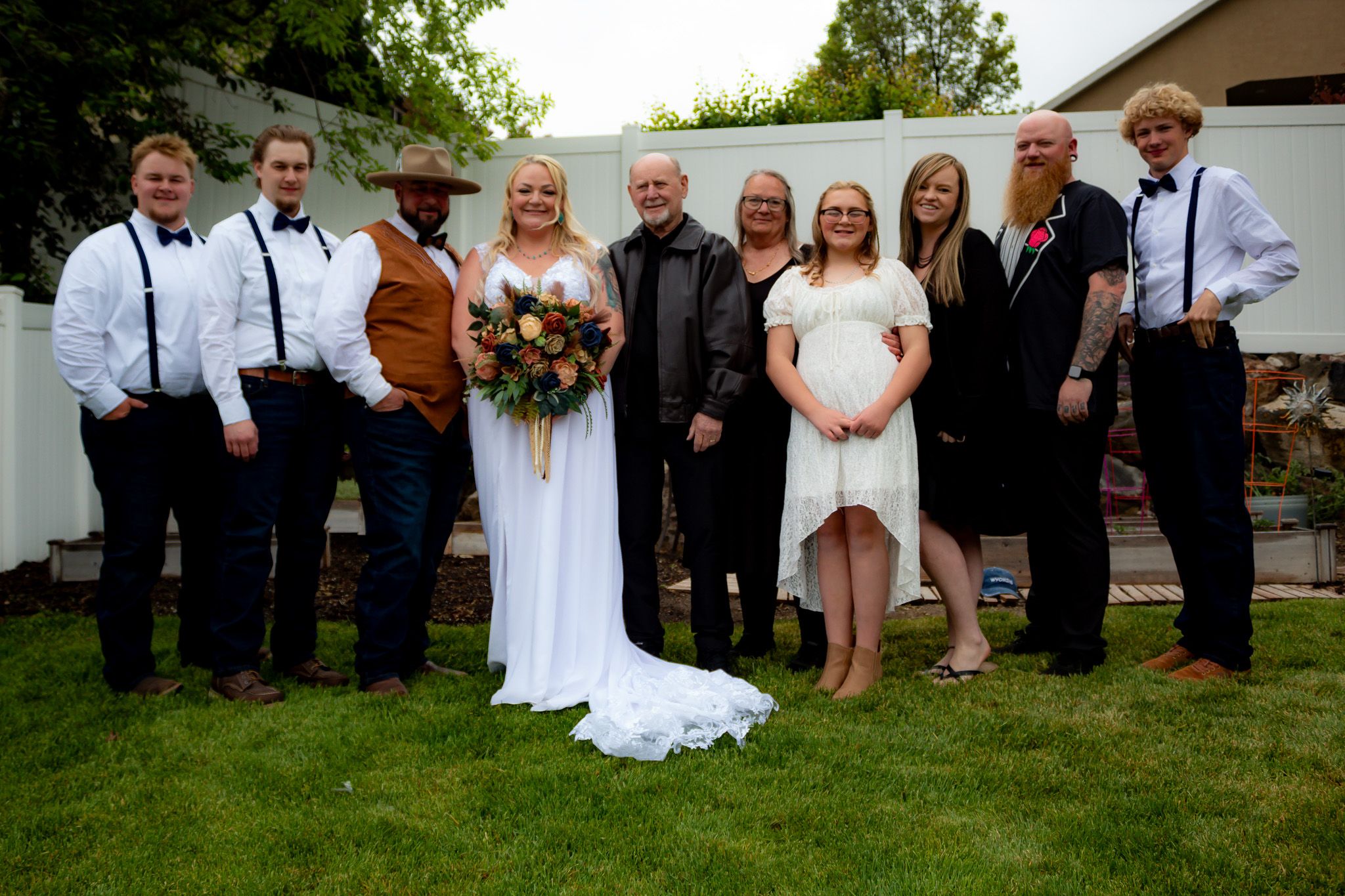 Wedding group photo showing formal attire with white fence and greenery in background.