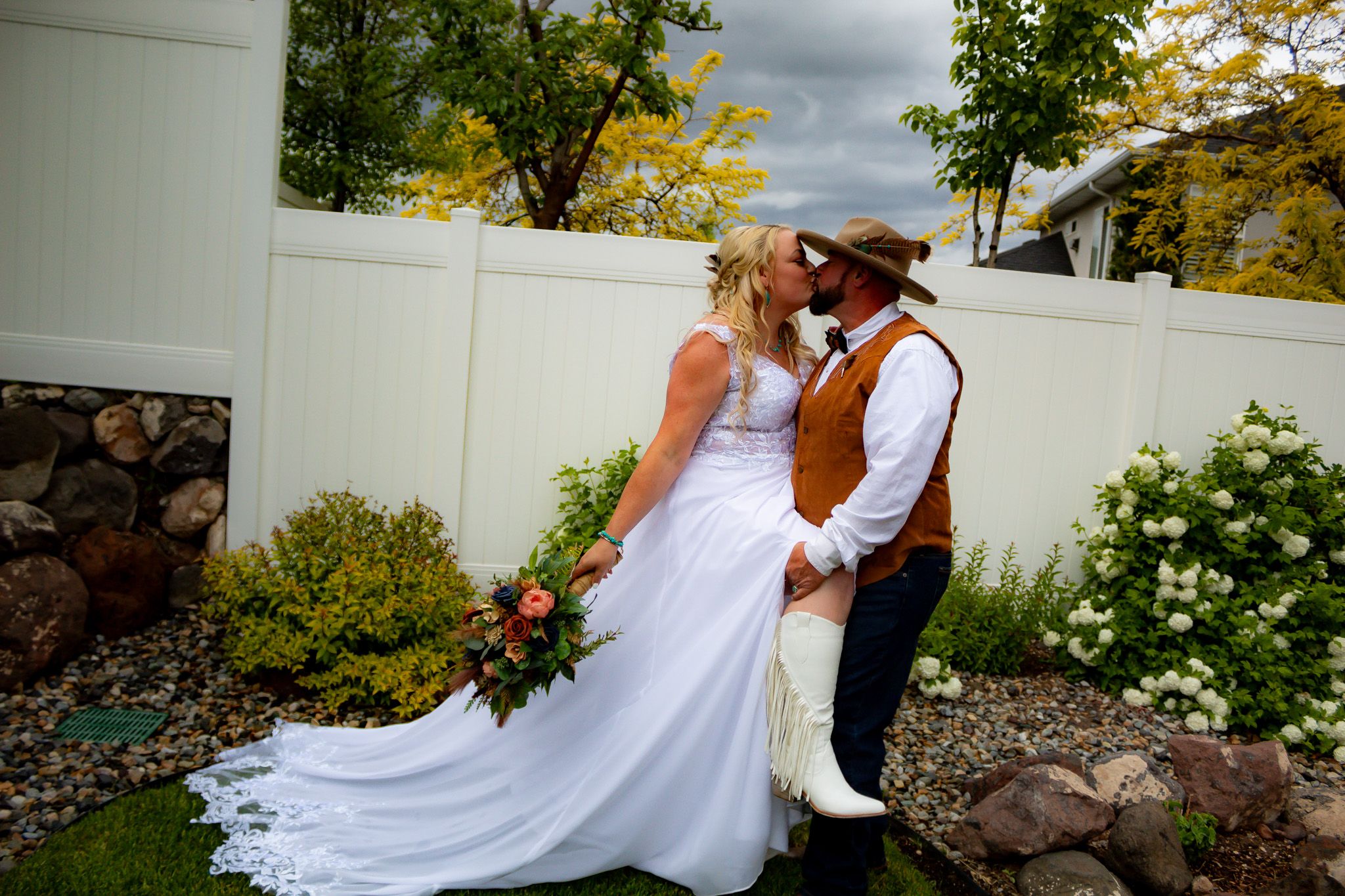 Couple in western wedding attire pose against white fence with flowing dress train.
