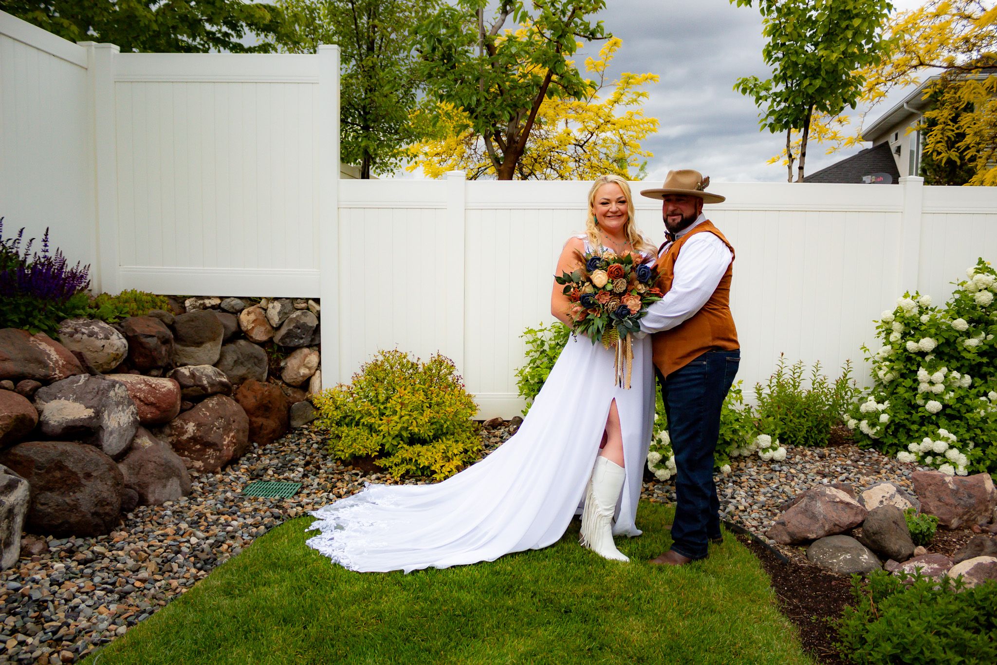 Wedding couple stands by stone wall and white fence in garden setting with autumn trees.