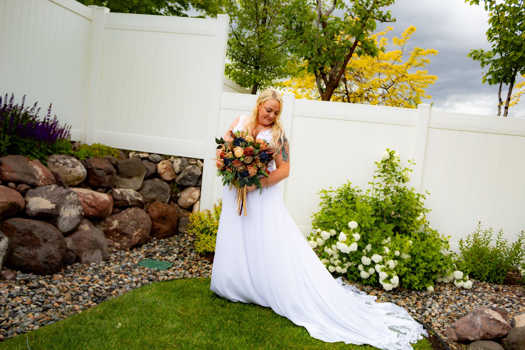 Bride in flowing white dress holds autumn bouquet in landscaped garden setting.